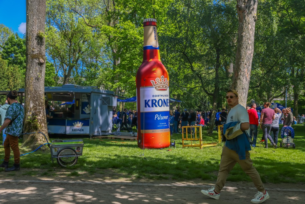 Eine große Flasche steht in einem belebten Park neben einem Verkaufsstand. Menschen gehen umher und genießen das sonnige Wetter.