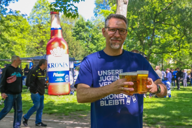 Ein Mann hält zwei Biergläser und steht vor einer großen Bierflasche im Park.