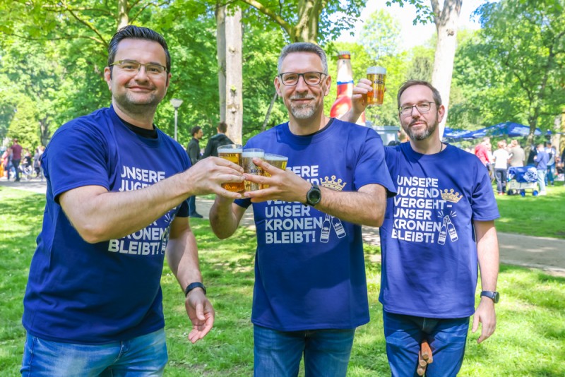 Drei Männer in blauen T-Shirts halten Biergläser in einem Park an.