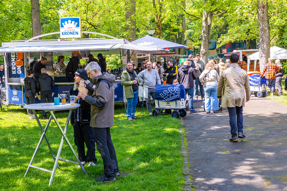 Mehrere Menschen stehen in einem Park  vor einem Dortmunder Kronen Ausschankwagen und feiern.