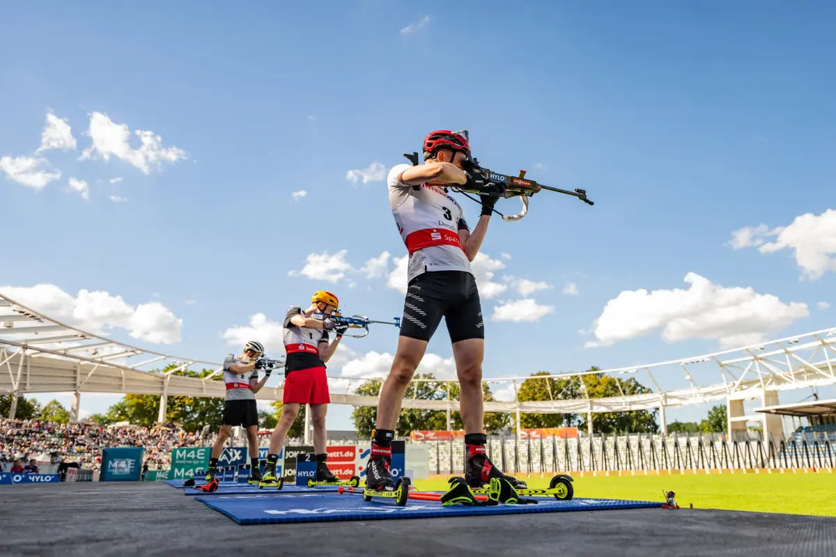 Drei Biathleten zielen mit Gewehren auf einer Rollski-Bahn. Sie tragen Helme und Sportkleidung unter blauem Himmel.